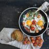 pan of fried eggs, bacon and cherry tomatoes with bread on dark table surface