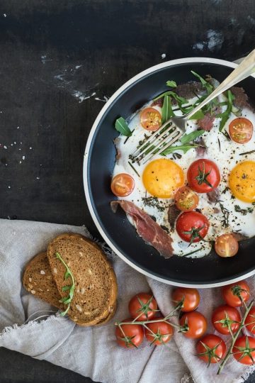 pan of fried eggs, bacon and cherry tomatoes with bread on dark table surface