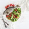 cooked sea bream fish with fresh vegetable salad on ceramic plate over white rustic wooden backdrop, top view