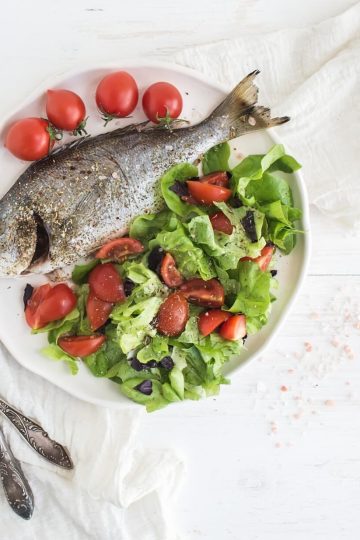 cooked sea bream fish with fresh vegetable salad on ceramic plate over white rustic wooden backdrop, top view