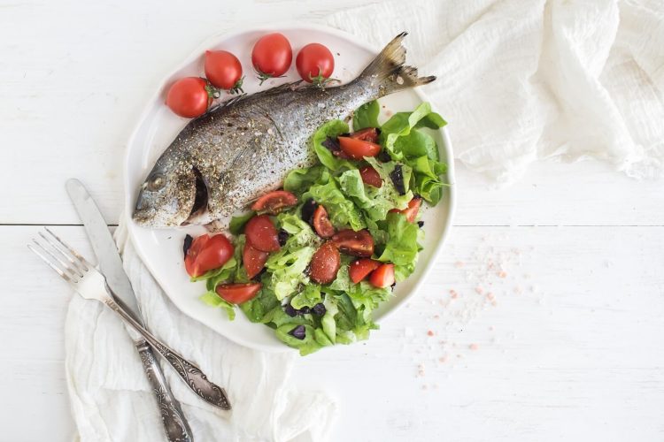 cooked sea bream fish with fresh vegetable salad on ceramic plate over white rustic wooden backdrop, top view