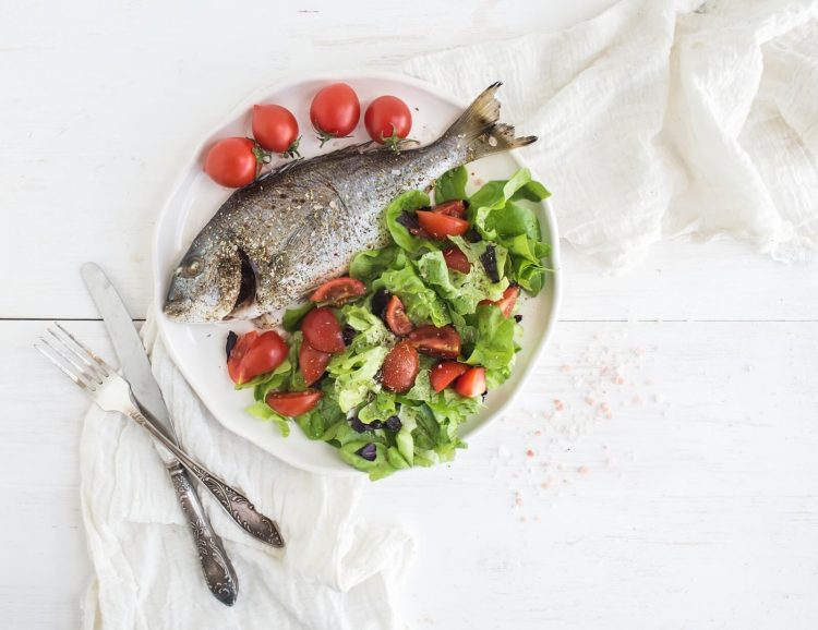 cooked sea bream fish with fresh vegetable salad on ceramic plate over white rustic wooden backdrop, top view